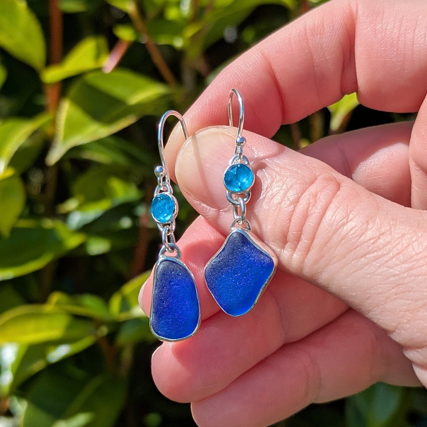 handholdingdanglyblueseaglassandgemstoneapatitesilverearringswithgreenplantbackground