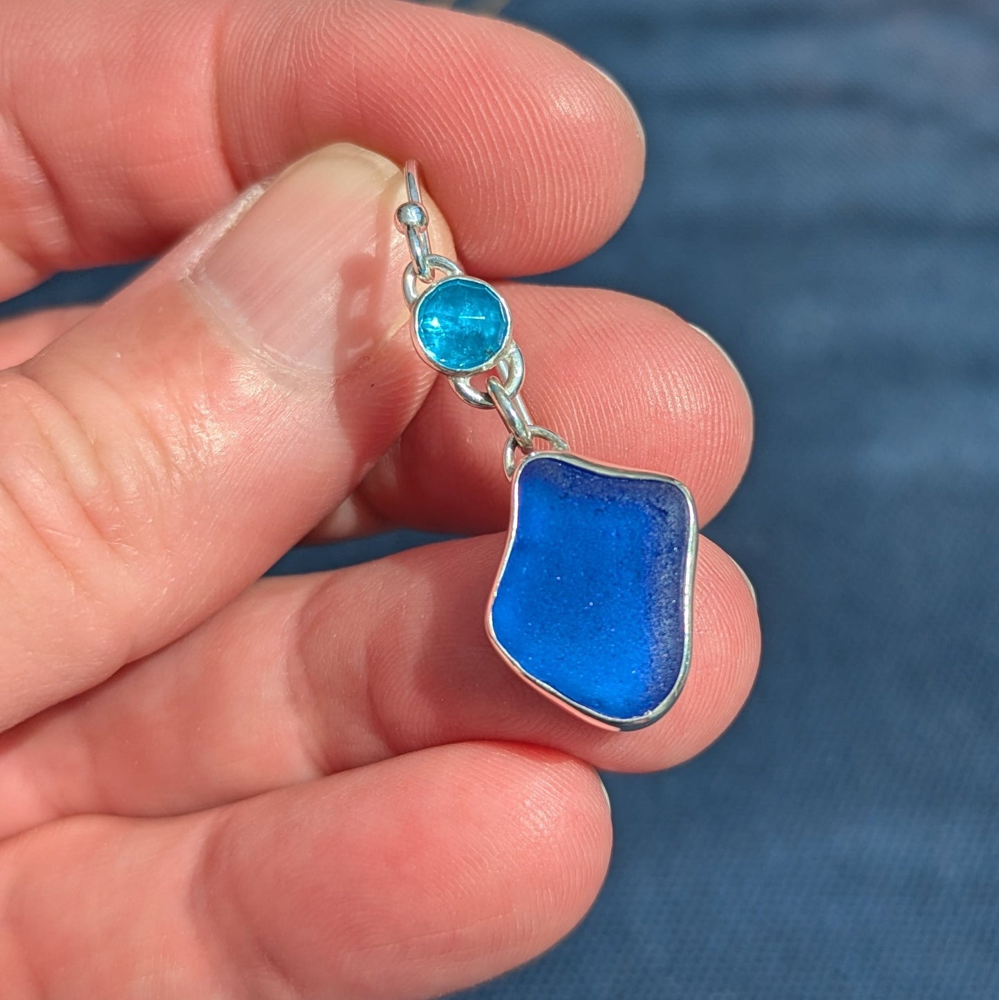 handholdingonedanglyblueseaglassandgemstoneapatitesilverearringwithbluebackground