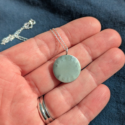 The back of a Recycled brushed silver round pendant, with the hand stamped words 'Ride the Tide' around a blue sea glass droplet.