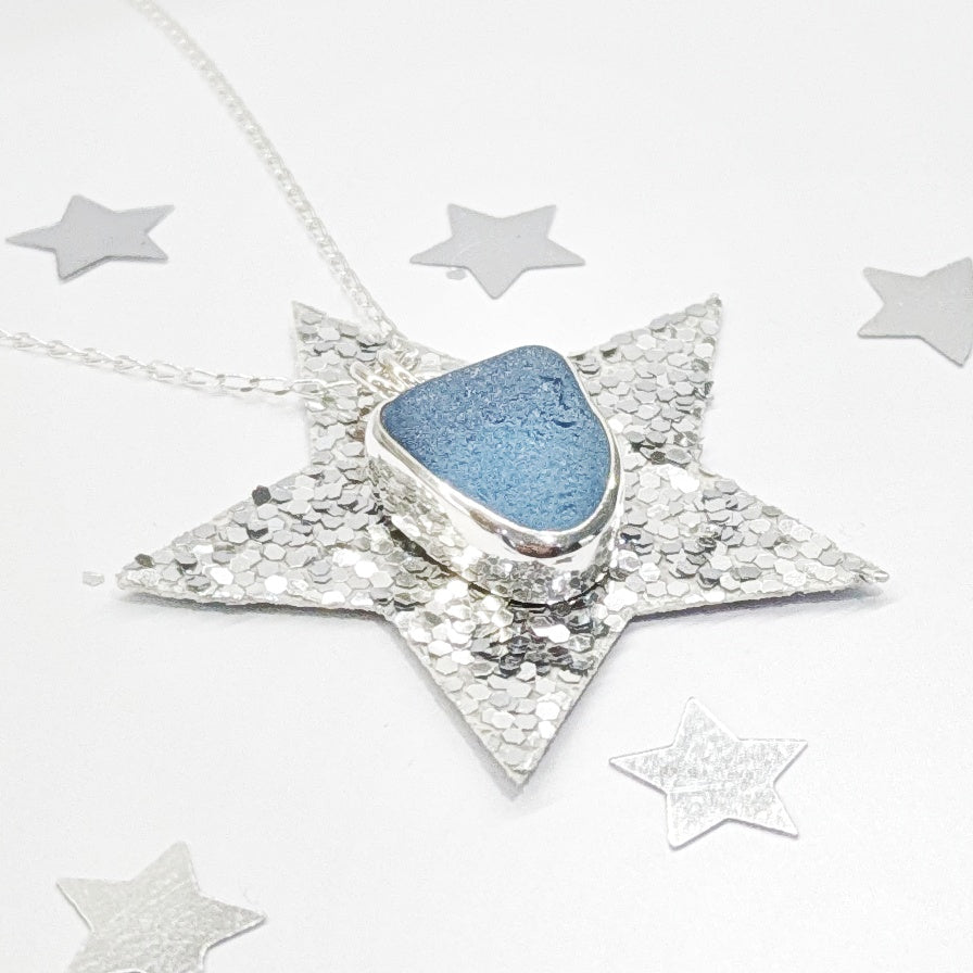 Cornish Blue sea glass silver pendant on silver chain with festive silver glitter stars in background.