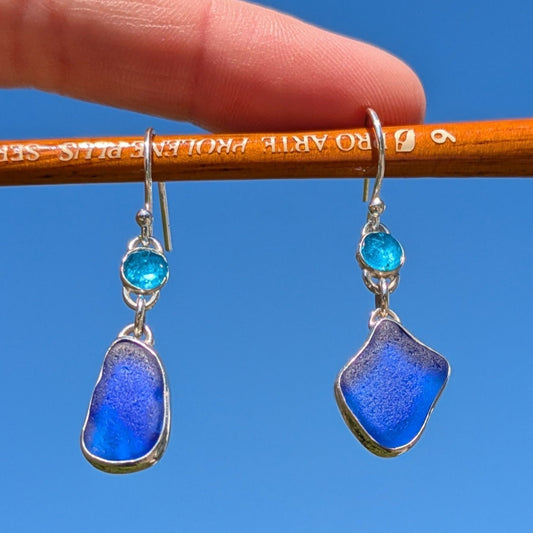 blue sea glass and neon blue gemstone apatite silver drop earrings  hanging from watercolour paint brush with blue sky background
