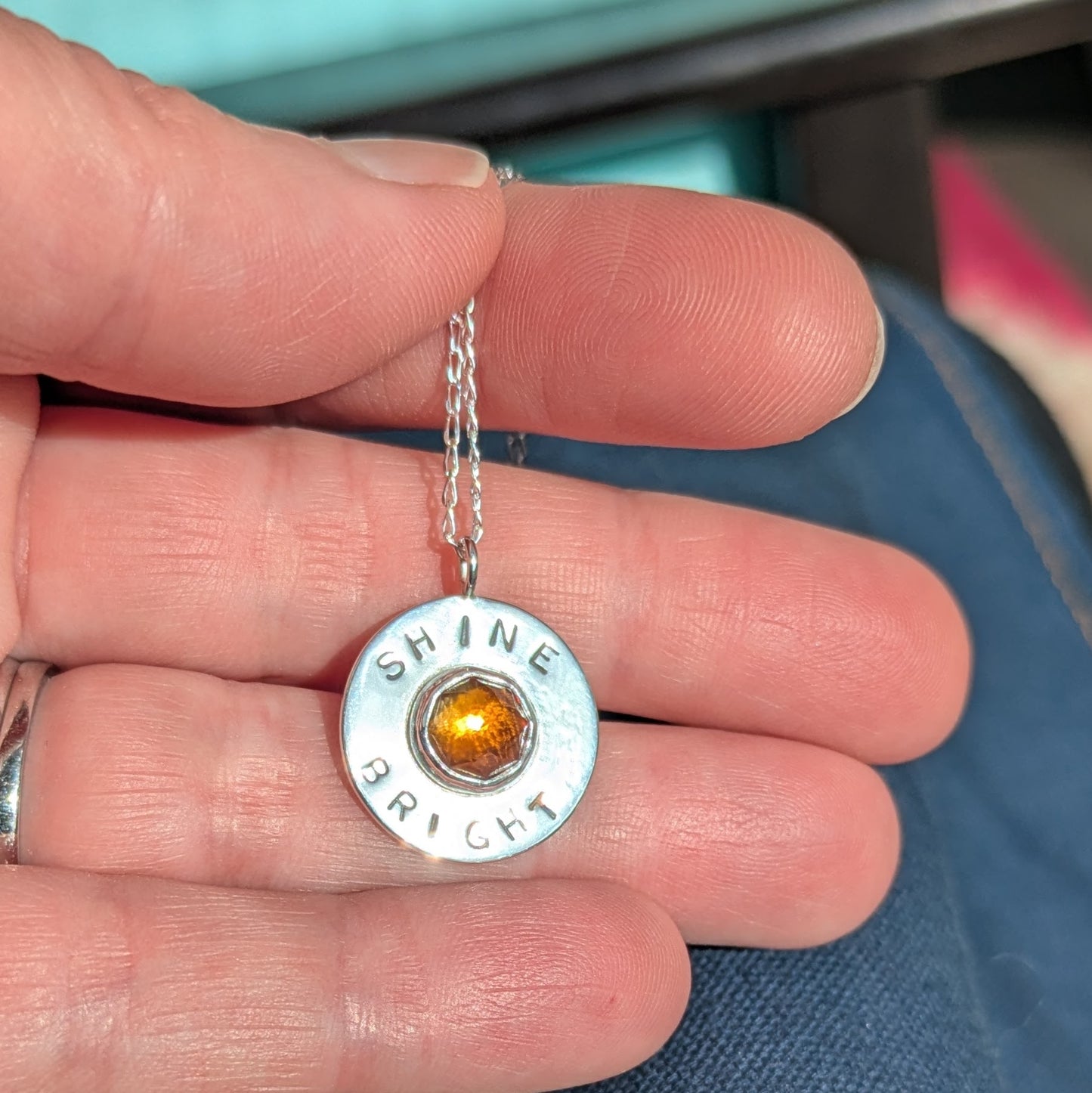 Handmade recycled silver circle pendant, featuring the words ' Shine Bright' around a central golden citrine gemstone.  Pictured in hand.
