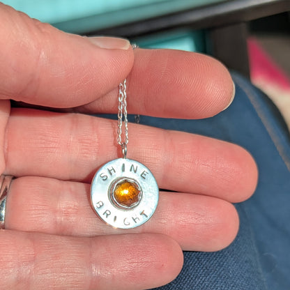 Handmade recycled silver circle pendant, featuring the words ' Shine Bright' around a central golden citrine gemstone.  Pictured in hand.