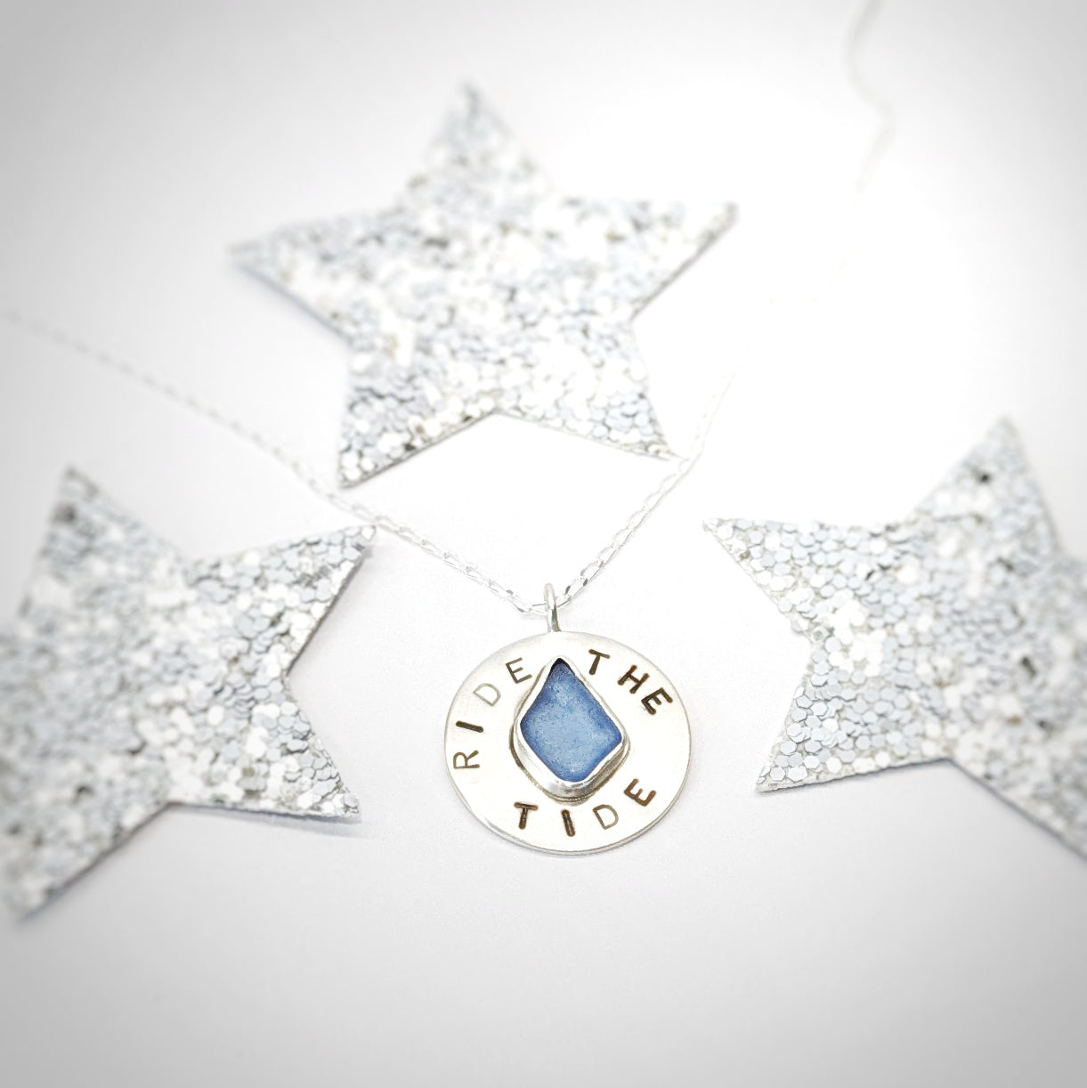 Recycled brushed silver round pendant, with the hand stamped words 'Ride the Tide' around a blue sea glass droplet, surrounded by festive silver glitter stars.