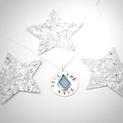 Recycled brushed silver round pendant, with the hand stamped words 'Ride the Tide' around a blue sea glass droplet, surrounded by festive silver glitter stars.