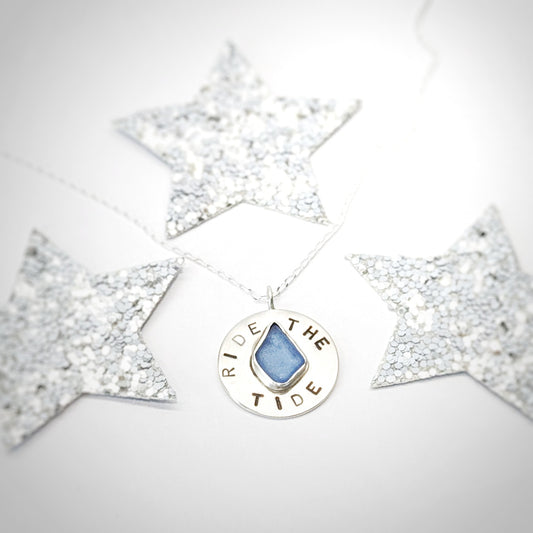 Recycled brushed silver round pendant, with the hand stamped words 'Ride the Tide' around a blue sea glass droplet, surrounded by festive silver glitter stars.
