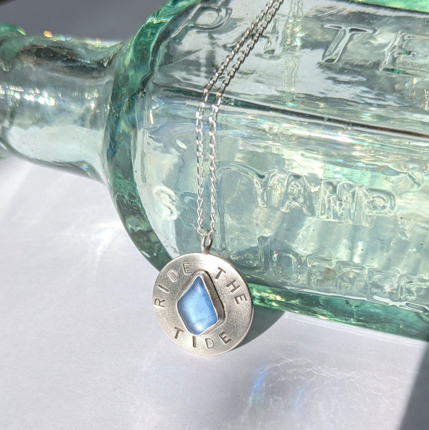 Recycled brushed silver round pendant, with the hand stamped words 'Ride the Tide' around a blue sea glass droplet.