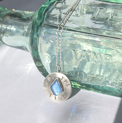 Recycled brushed silver round pendant, with the hand stamped words 'Ride the Tide' around a blue sea glass droplet.