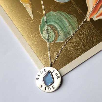 Recycled brushed silver round pendant, with the hand stamped words 'Ride the Tide' around a blue sea glass droplet. Pictured sitting next to a gold sea shell card.
