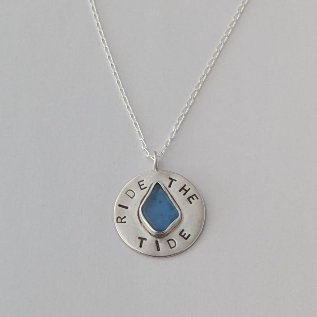 Recycled brushed silver round pendant, with the hand stamped words 'Ride the Tide' around a blue sea glass droplet. Pictured with chain on a white background.