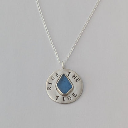 Recycled brushed silver round pendant, with the hand stamped words 'Ride the Tide' around a blue sea glass droplet. Pictured with chain on a white background.