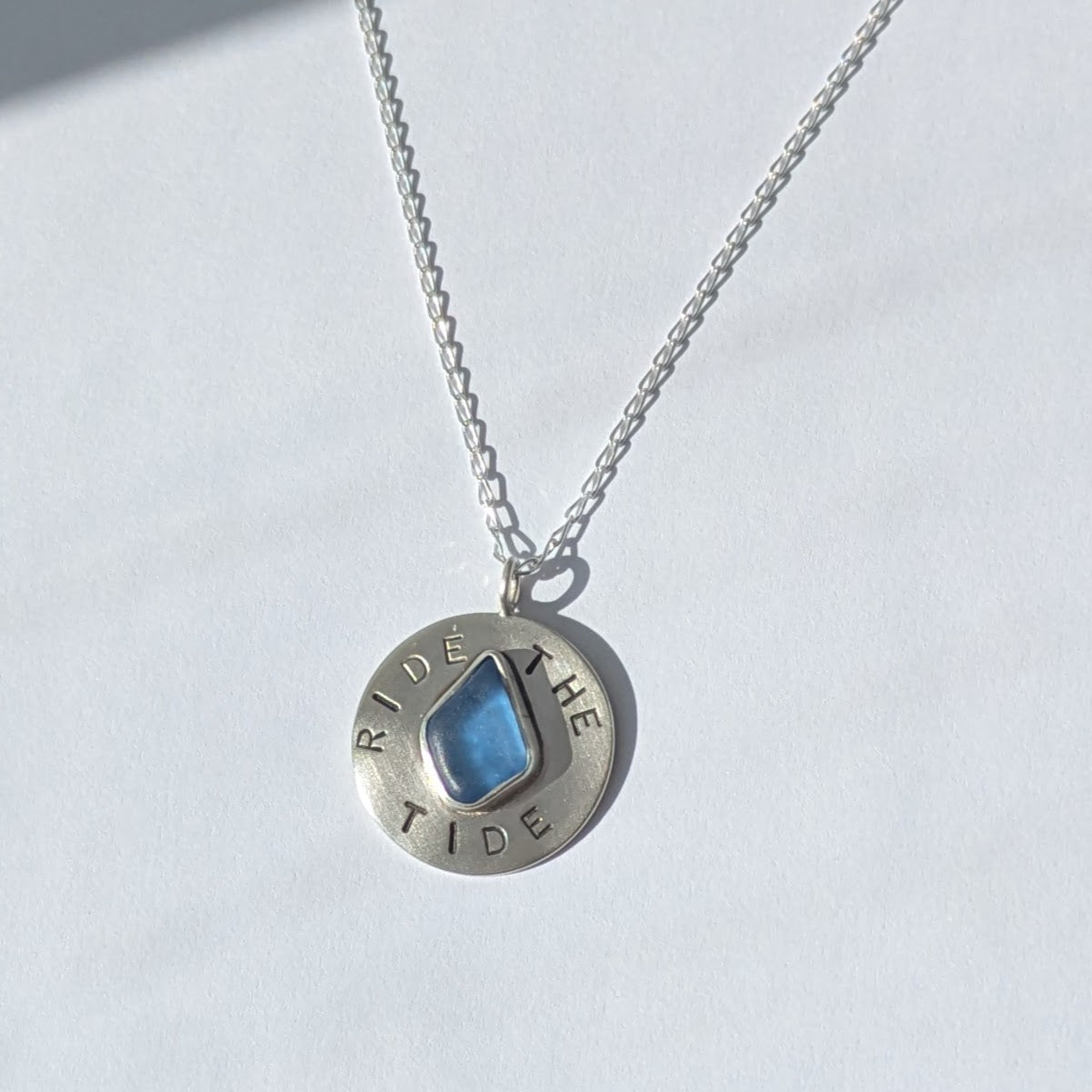 Recycled brushed silver round pendant, with the hand stamped words 'Ride the Tide' around a blue sea glass droplet. Pictured on a chain with white background.