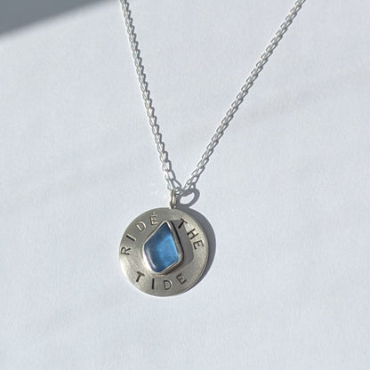 Recycled brushed silver round pendant, with the hand stamped words 'Ride the Tide' around a blue sea glass droplet. Pictured on a chain with white background.
