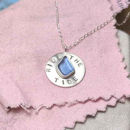 Recycled brushed silver round pendant, with the hand stamped words 'Ride the Tide' around a blue sea glass droplet. Pictured on a pink polishing cloth.