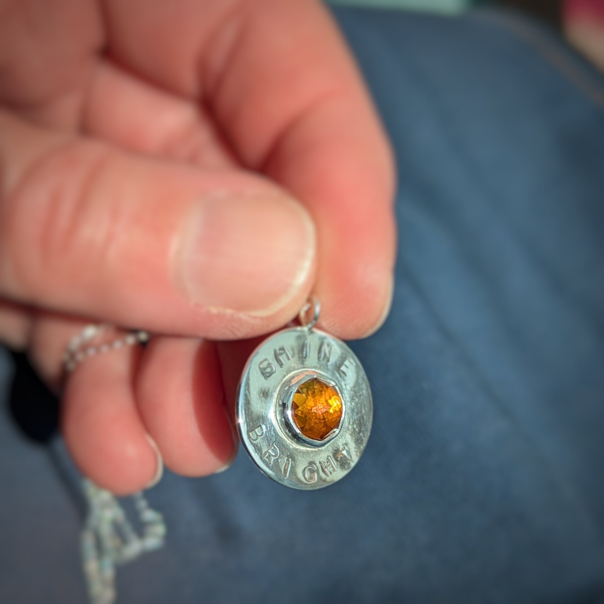 Handmade recycled silver circle pendant, featuring the words ' Shine Bright' around a central golden citrine gemstone.  Pictured in hand.