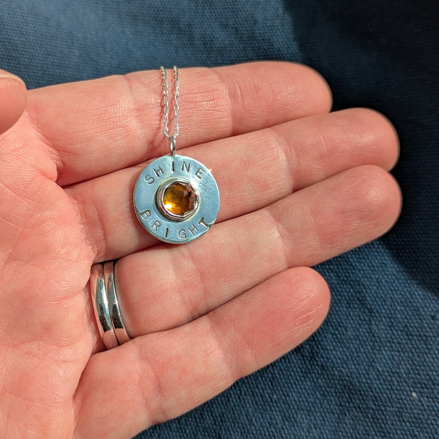Handmade recycled silver circle pendant, featuring the words ' Shine Bright' around a central golden citrine gemstone.  Pictured in hand.