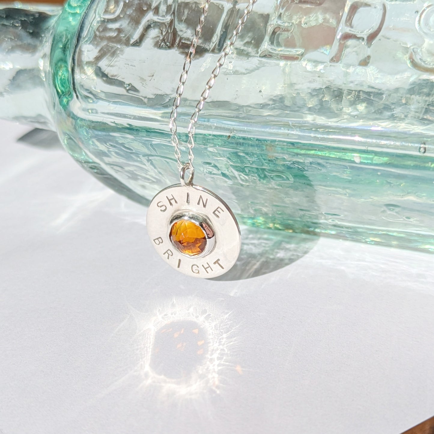 Handmade recycled silver circle pendant, featuring the words ' Shine Bright' around a central golden citrine gemstone. Pictured leaning against an old glass bottle in the sunshine.
