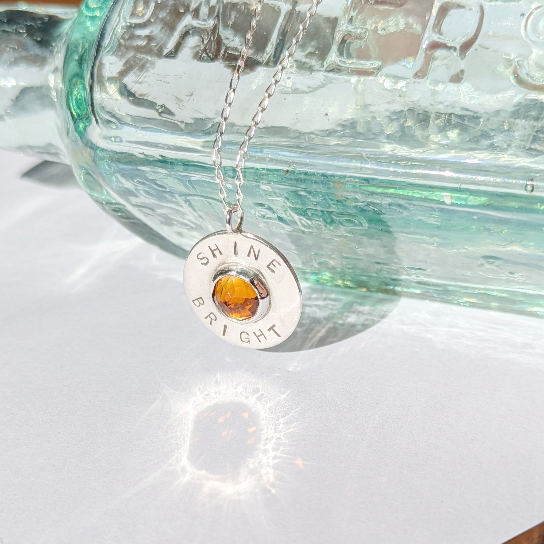 Handmade recycled silver circle pendant, featuring the words ' Shine Bright' around a central golden citrine gemstone. Pictured leaning against an old glass bottle in the sunshine.