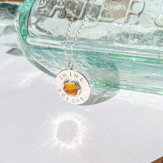 Handmade recycled silver circle pendant, featuring the words ' Shine Bright' around a central golden citrine gemstone. Pictured leaning against an old glass bottle in the sunshine.