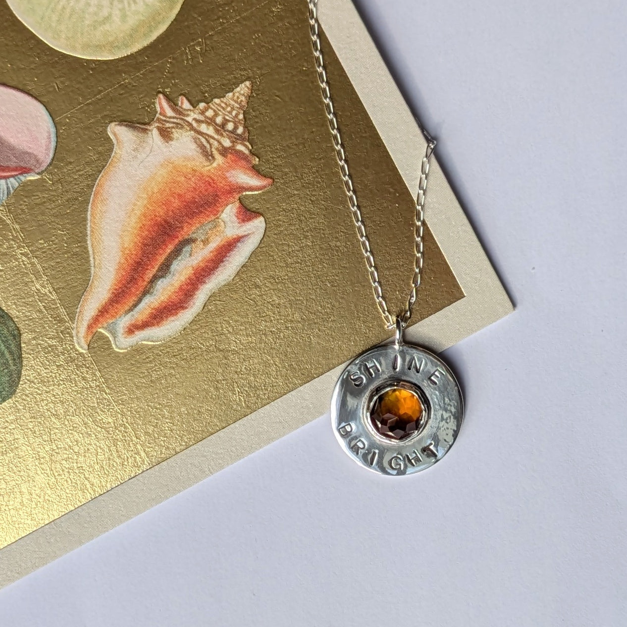 Handmade recycled silver circle pendant, featuring the words ' Shine Bright' around a central golden citrine gemstone.  Pictured leaning against a gold shell card.