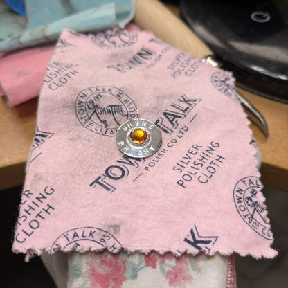 Handmade recycled silver circle pendant, featuring the words ' Shine Bright' around a central golden citrine gemstone.  Pictured on a pink polishing cloth.