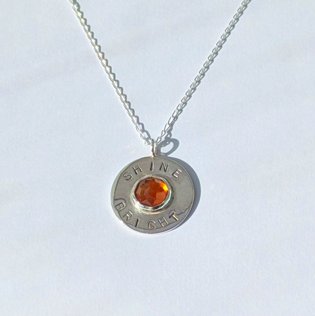 Handmade recycled silver circle pendant, featuring the words ' Shine Bright' around a central golden citrine gemstone.  Pictured on white background.