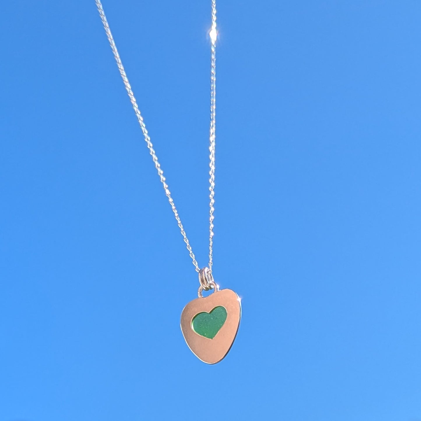 Green Sea Glass silver pendant with heart shape cut out, with silver chain held up to blue sky.