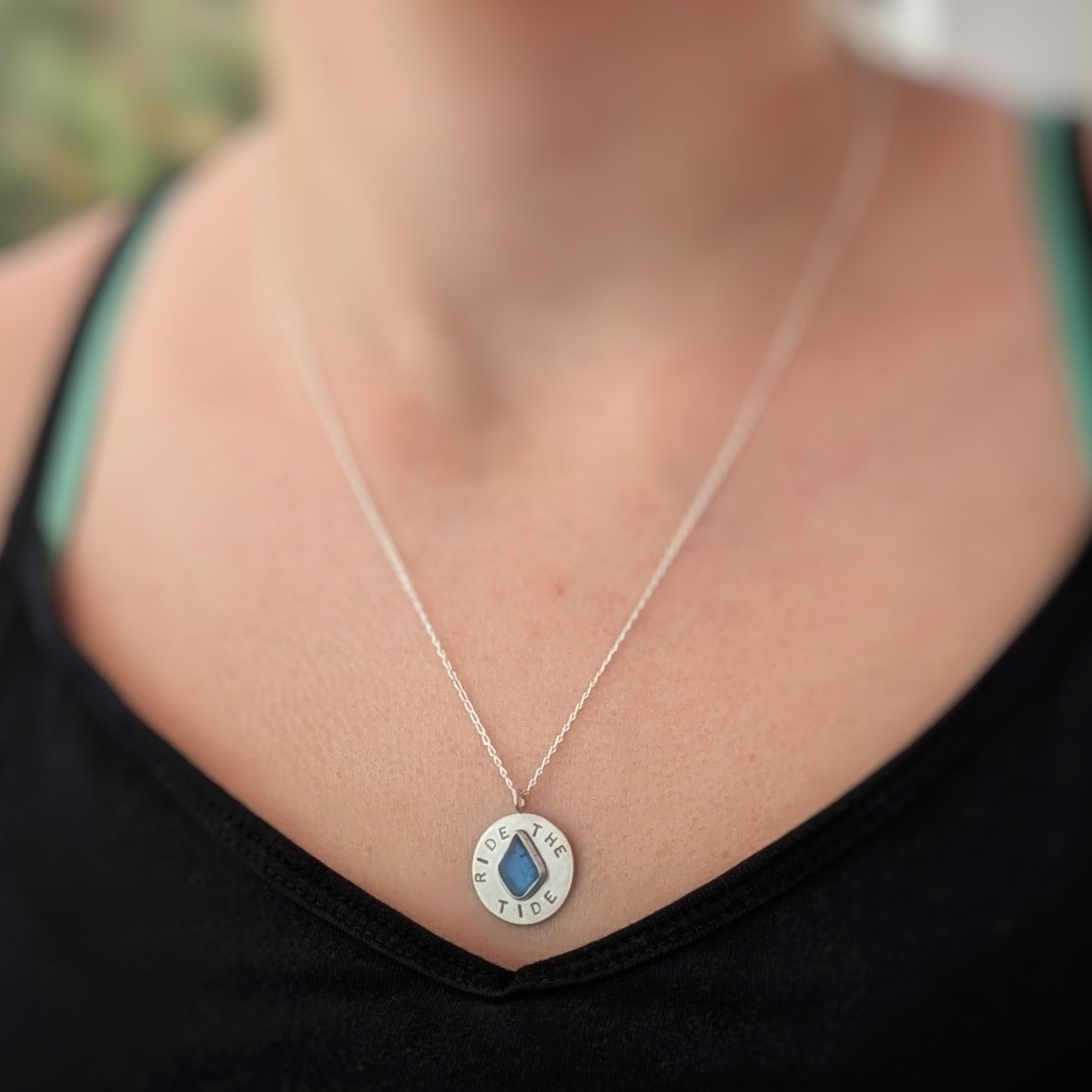 Recycled brushed silver round pendant, with the hand stamped words 'Ride the Tide' around a blue sea glass droplet. Pictured worn by a model.
