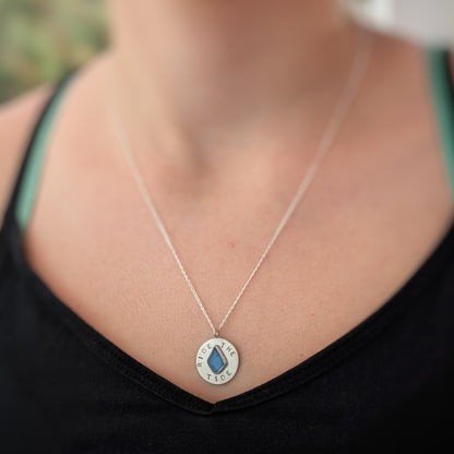 Recycled brushed silver round pendant, with the hand stamped words 'Ride the Tide' around a blue sea glass droplet. Pictured worn by a model.