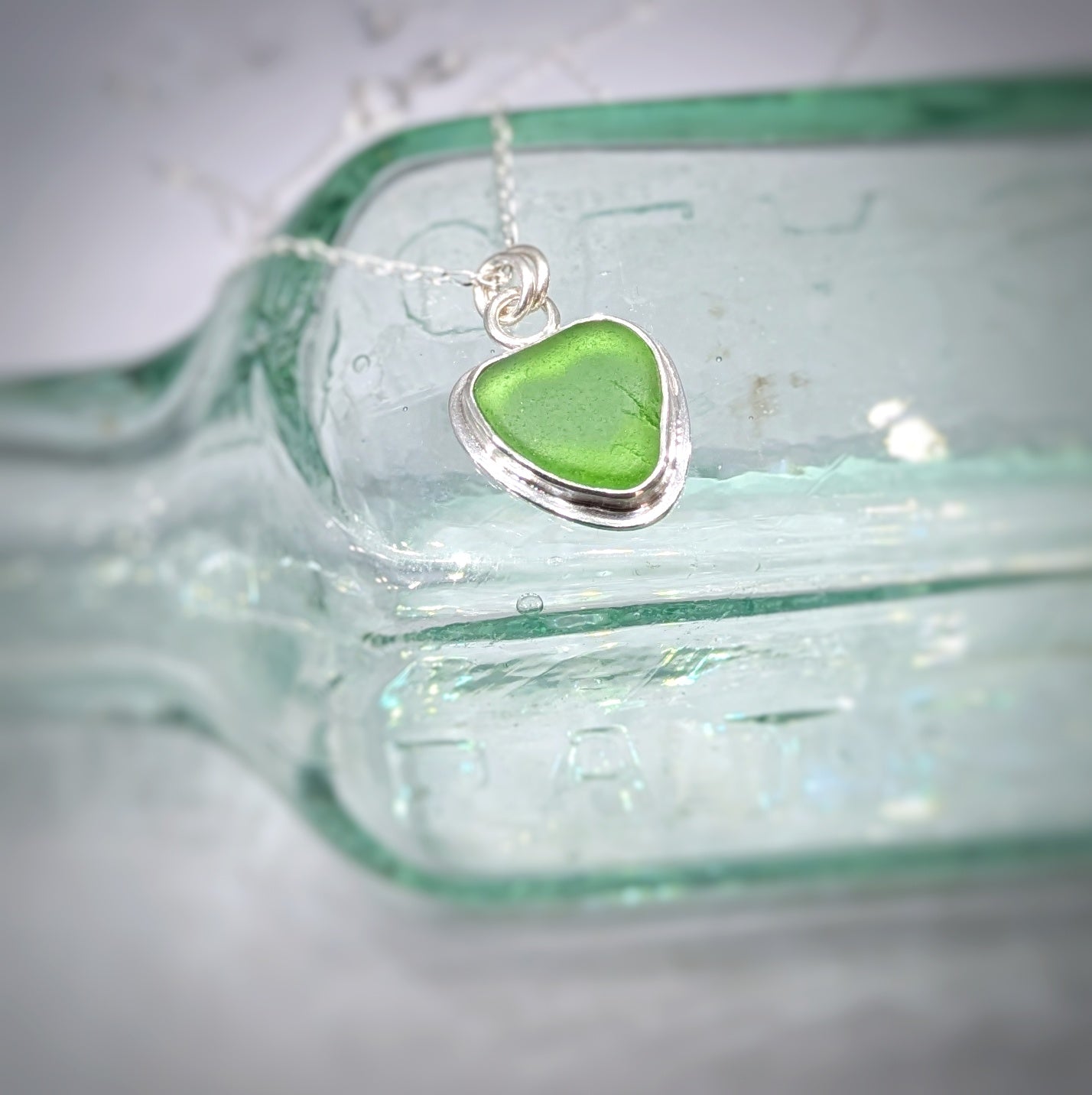 Green Sea Glass silver pendant with heart shape cut out, with silver chain sitting on old glass bottle.