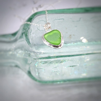 Green Sea Glass silver pendant with heart shape cut out, with silver chain sitting on old glass bottle.