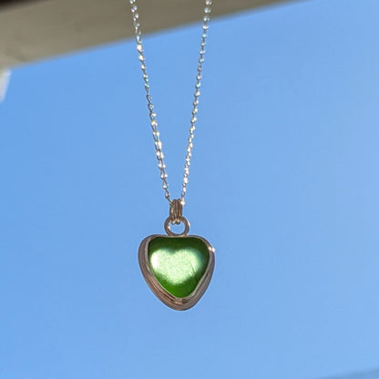 Green Sea Glass silver pendant with heart shape cut out, with silver chain held up to blue sky with light shining through heart cut out.