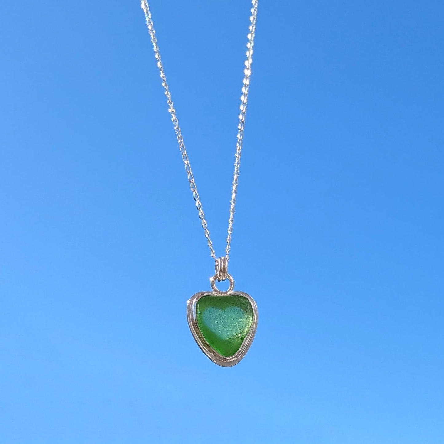 Front view of Green Sea Glass silver pendant with heart shape cut out, with silver chain held up to blue sky.