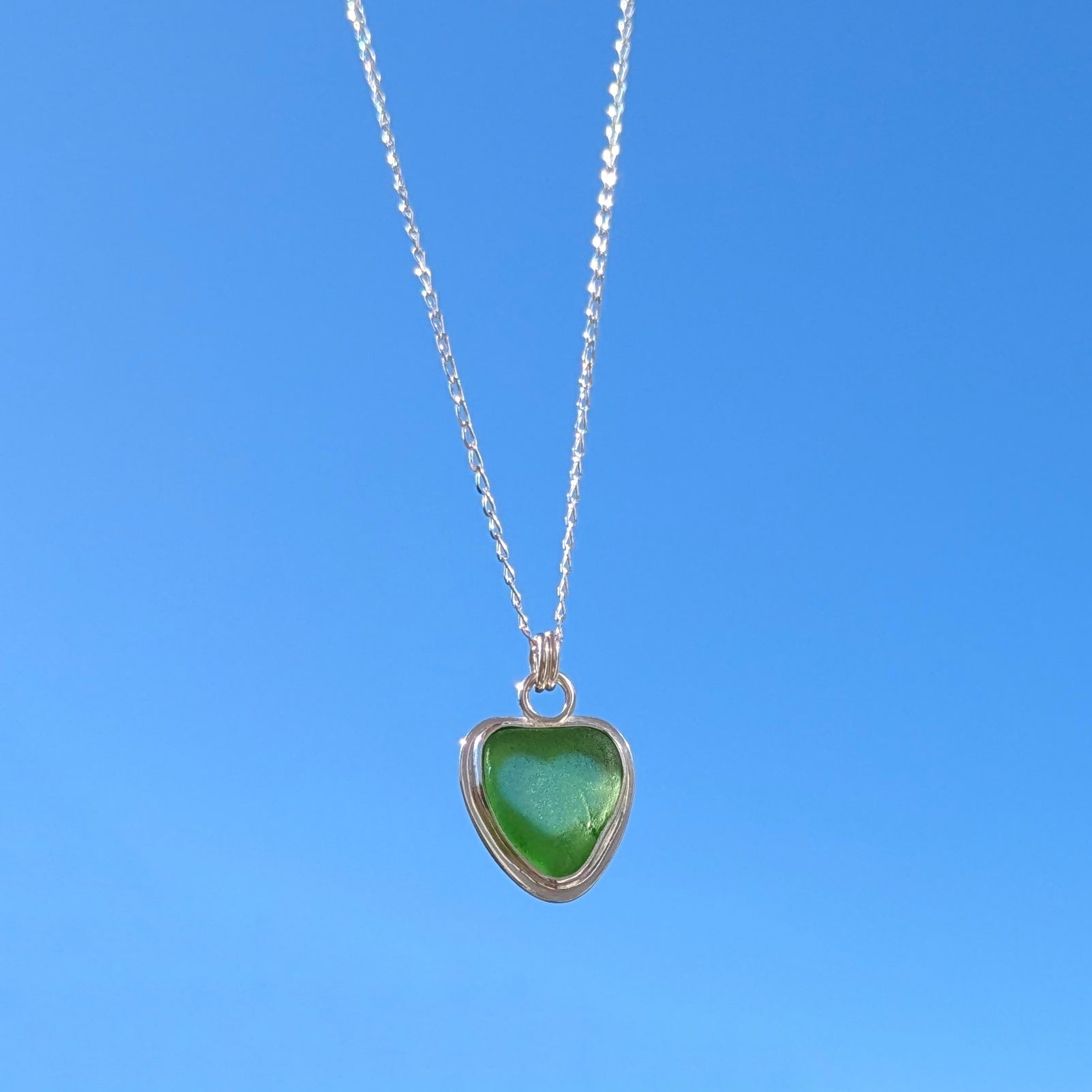Front view of Green Sea Glass silver pendant with heart shape cut out, with silver chain held up to blue sky.