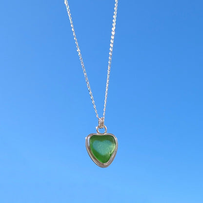 Front view of Green Sea Glass silver pendant with heart shape cut out, with silver chain held up to blue sky.