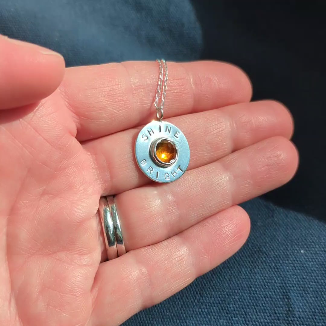 Handmade recycled silver circle pendant, featuring the words ' Shine Bright' around a central golden citrine gemstone.  Pictured in hand.