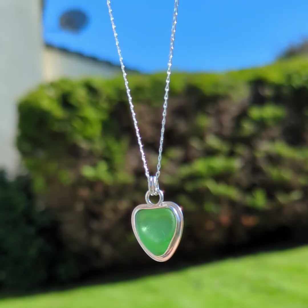 Video of Green Sea Glass silver pendant with heart shape cut out, with silver chain hanging up with blue sky & greenery background.