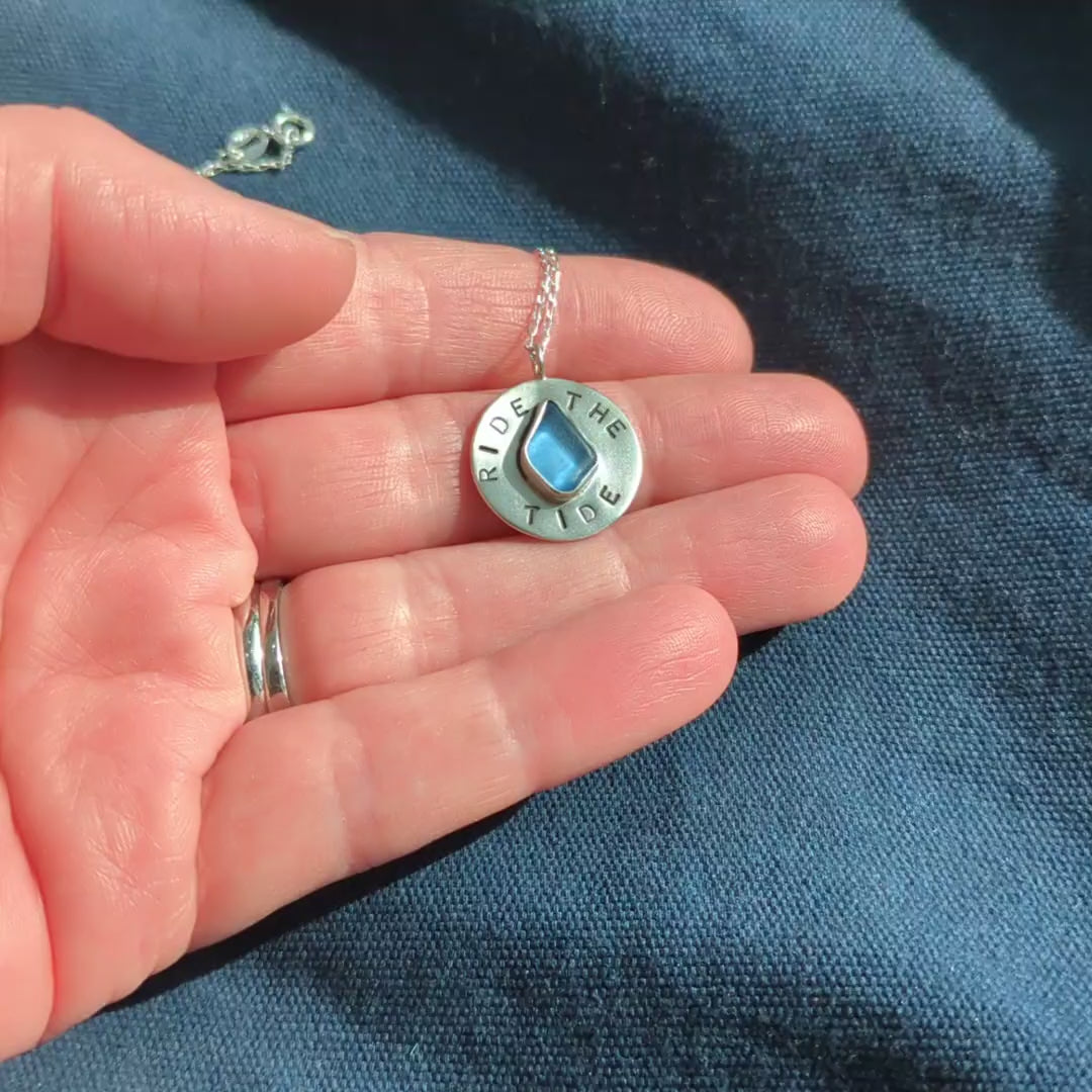 Recycled brushed silver round pendant, with the hand stamped words 'Ride the Tide' around a blue sea glass droplet. Video shows pendant in hand.