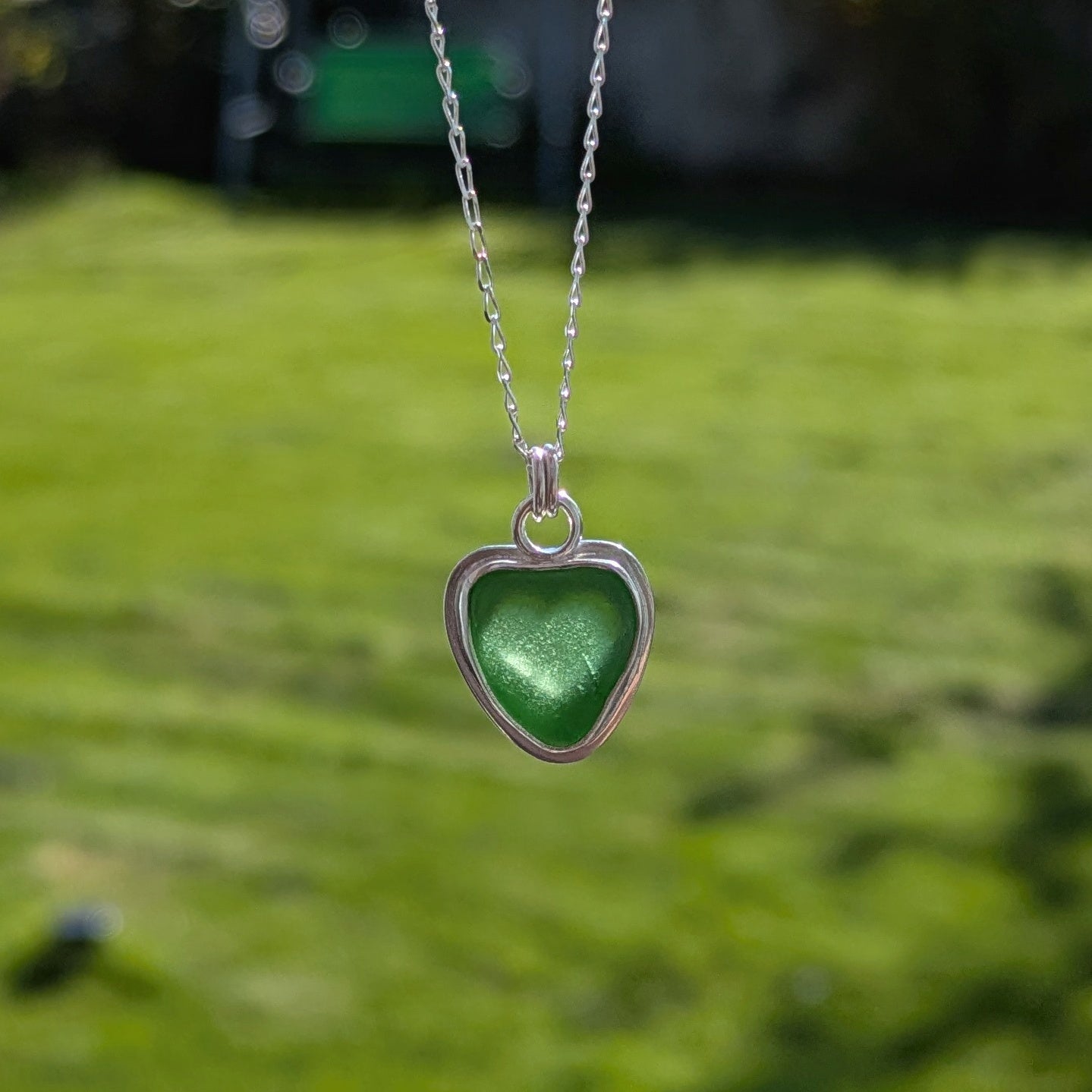 Green Sea Glass silver pendant with heart shape cut out, with silver chain held up with grass background and sun shining through heart cut out.