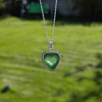 Green Sea Glass silver pendant with heart shape cut out, with silver chain held up with grass background and sun shining through heart cut out.